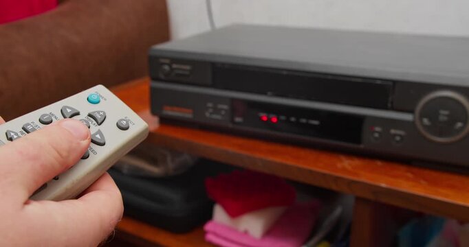 Hand presses eject button on remote control as VHS tape slides out of video cassette recorder. Retro media device and pre-digital home entertainment technology