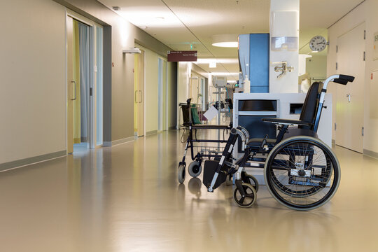 A wheelchair is seen in a corridor of a modern Dutch hospital. The hallway has light colors and doors leading to different rooms. The setting is bright and quiet, showcasing a healthcare space.