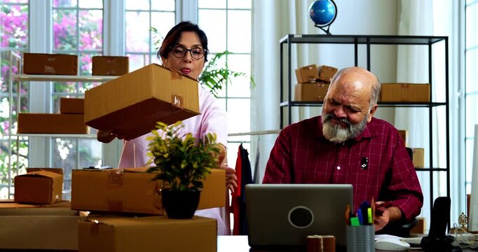 Indian senior couple celebrating success in home based E-commerce business while managing online orders, dispatching parcels, packing cardboard boxes and working together in home office