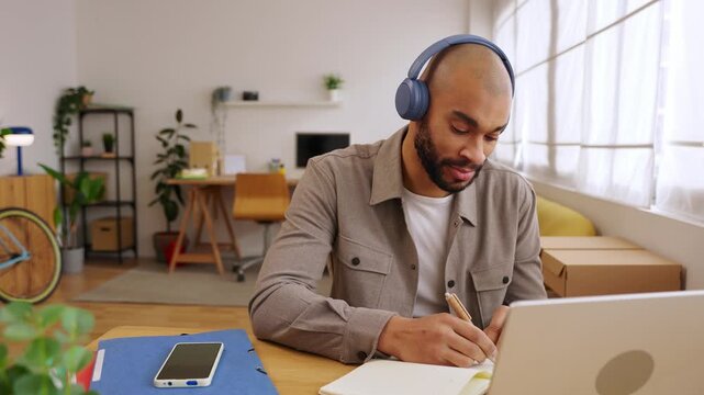 Young adult African American man taking notes during online course. Adult student learning from laptop while writing in notebook at home. Education and e-learning concept. 