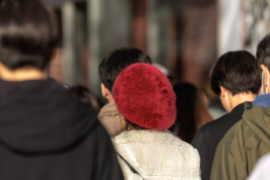 Commuters walking in soft warm sunlight with focus on fuzzy crimson beret of woman wearing cream colored teddy‑style coat in urban setting.