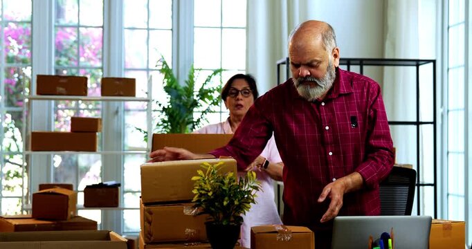 Indian senior couple making products ready to be dispatched, while doing E-commerce business from home managing online orders, packing cardboard boxes, counting garments and preparing parcels