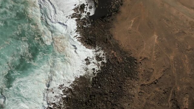 Aerial view of a coastline where turquoise waves crash against dark, rocky shores leading to dry, sandy earth.