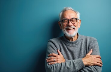 Obraz premium Happy elder man with glasses smiles broadly, arms crossed against blue background. Mature person expresses joy and contentment, embodying peaceful retirement. He looks relaxed and full of life.