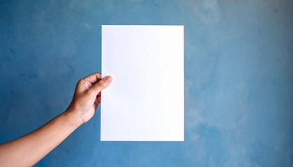 Obraz premium Hand holding a blank white paper against a textured blue wall for design mockups and presentations.