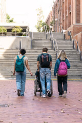 Three diverse students walking on campus, one pushing a friend in a wheelchair, promoting...