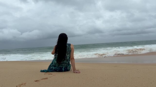 Woman sitting on a sandy beach watching a stormy Indian Ocean in Sri Lanka. Moody overcast weather, waves crashing, and solitude. Mindfulness and nature concept.