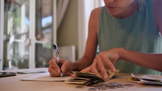 Woman at home counting cash and coins at a table, writing notes with a pen while tracking expenses and budgeting household finances with focused concentration
