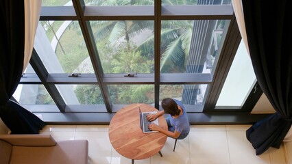 Woman working remotely on a laptop at a wooden table in front of a grand window, embracing...