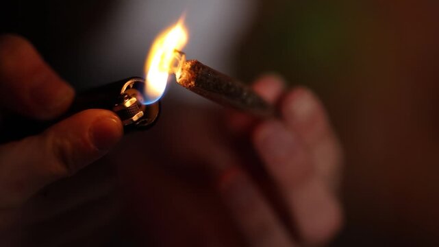 Close-up view of male hands lighting a handmade cannabis cigarette in a dark environment