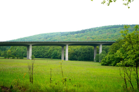 A long concrete highway bridge spans across a wide green meadow landscape. Massive grey support pillars hold the elevated road structure firm. A forested green hill rises in the background.