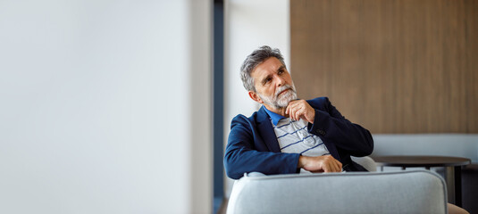 Male professional with hand on chin looking away while sitting on sofa in office contemplating...