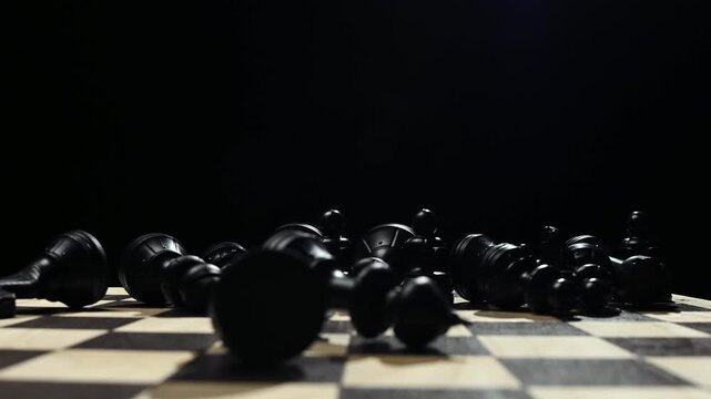 Black chess pieces on a board stand in a row. A man's hand knocks down the pieces on a chessboard on a black background 4к