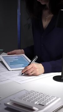 Woman working late night in office, multitasking with a tablet and writing on documents, managing heavy paperwork load. Audit and taxes in business