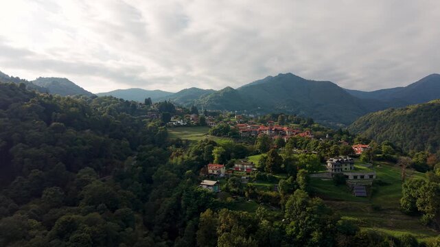 Drone view of Alzo, a small village nestled in green hills and forests with mountains in the background in Piedmont, Italy. Aerial forward, copy space