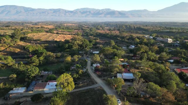 White van navigating winding dirt road in rural Mexico at dawn. Aerial view shows low mist across valley floor between sunlit hills, flowering trees, and small village clusters.