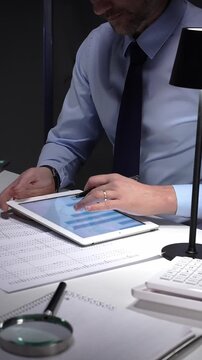 Businessman working late analyzing financial data at office, holding tablet with charts, writing notes on documents. Audit and taxes in business