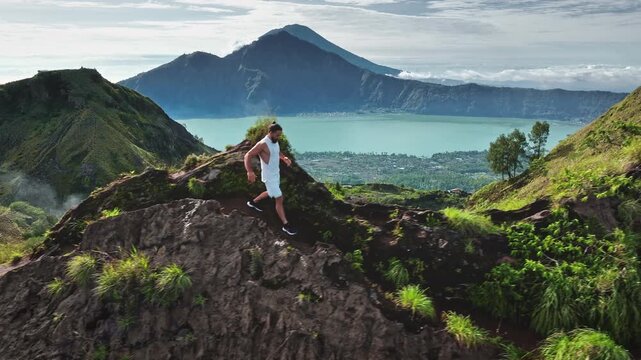Man running uphill on a rugged Batur mountain ridge path, enjoying the expansive panoramic view of a majestic volcano and a vibrant turquoise lake under a bright sky in Bali, Indonesia. Drone shot