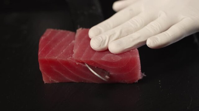 Professional chef with gloves slicing raw tuna fillet with a sharp knife on a black cutting board for preparing delicious sushi, sashimi, or poke bowl in a restaurant kitchen