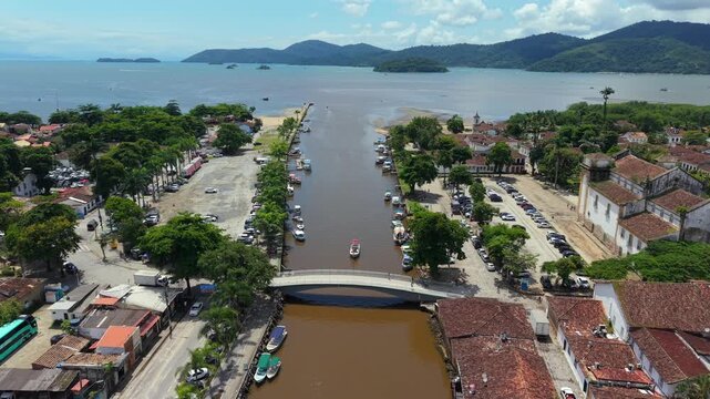 Paraty Centro Hist&oacute;rico in Rio de Janeiro state, Brazil, showing colonial waterfront channel leading to Ba&iacute;a de Paraty with church facade, palm lined promenade and anchored boat, drone pulling out