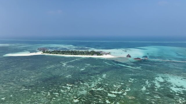 Distant aerial drone shot featuring a left-orbiting movement that reveals the massive scale of turquoise shallows surrounding a lush tropical island with beach villas in the Maldives