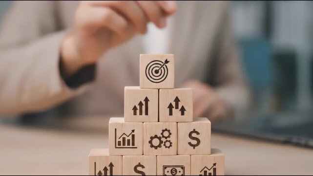 Close-up of a hand placing a wooden block with a target icon on top of stacked cubes displaying business symbols, subtle alignment motion with slow cinematic push-in, soft neutral lighting and shallow
