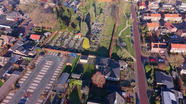 High angle aerial view of a neat European cemetery in a residential neighborhood. Features organized gravestones, lush greenery, red roofed houses, a parking lot, and a small park under soft sunlight.