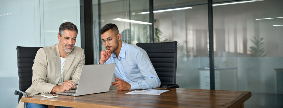 Two busy professional business men working together talking using laptop in office. Male manager and employee looking at computer discussing investment business project analysis at office meeting.