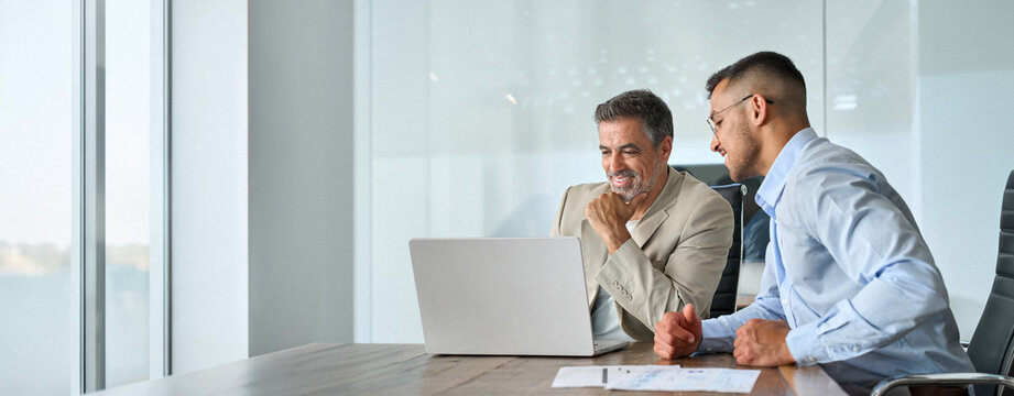 Happy busy professional team of two business men executive having discussion, male partners working using laptop looking at computer analyzing online tech data at work in office sitting at desk.