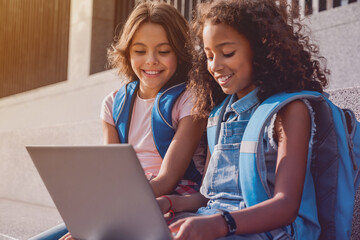 Two happy elementary middle school girls friends pupils classmates using laptop gadgets looking on screen doing homework project together sitting on the stairs steps outdoors © InsideCreativeHouse
