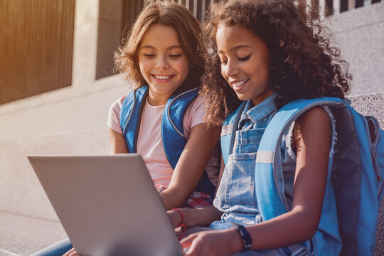 Two happy elementary middle school girls friends pupils classmates using laptop gadgets looking on screen doing homework project together sitting on the stairs steps outdoors