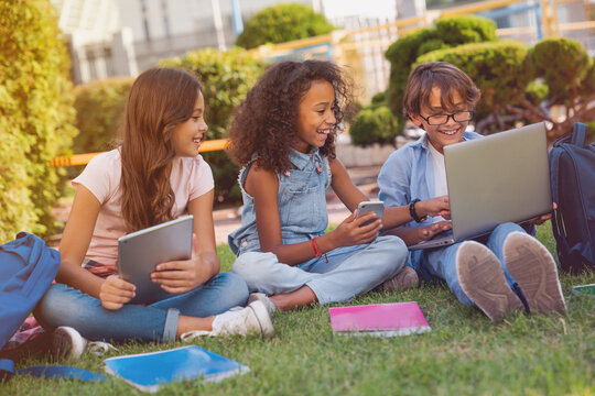 Happy little school children elementary middle school pupils schoolmates with digital gadgets laptop tablet smart phone backpacks notebooks sitting at the grass park outdoors doing homework