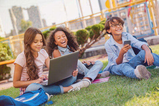 Group of smiling multiethnic elementary middle school children pupils classmates sitting on playground grass looking at laptop computer screen doing homework together