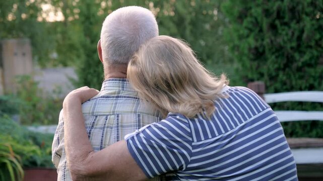 Elderly caucasian couple hugs while sitting on bench in backyard garden at golden hour. Woman puts her head on man shoulder as sign of love and care. Back view old couple in park. Visit sick relatives