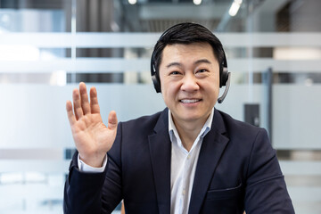 Asian businessman wearing a headset and smiling. Waving his hand hello or goodbye during a remote...