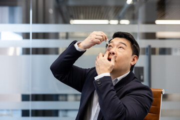 Asian man in business attire at office desk applying eye drops to his irritated eyes, managing daily eye irritation and practicing self-care while working