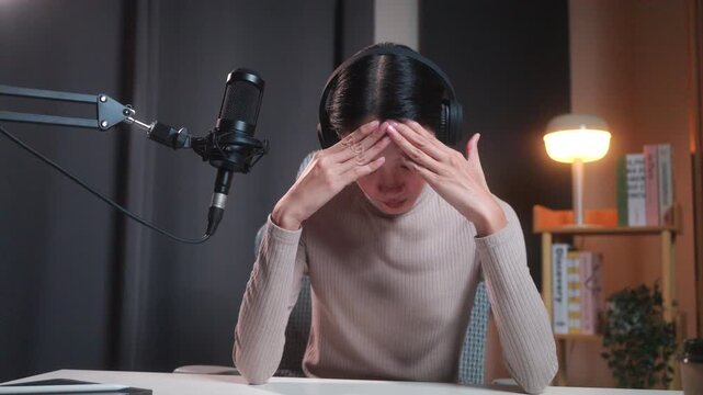 Frustrated young asian female content creator experiencing a creative block or technical issue during a podcast recording session, sitting at a desk with microphone and digital tablet.