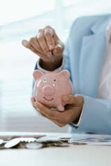 Businesswoman placing a coin into a pink piggy bank on office desk with calculator and charts,...
