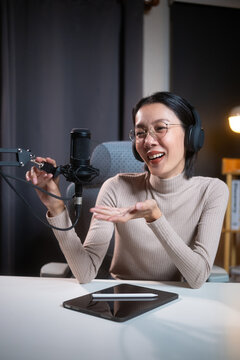 Smiling beautiful asian female podcaster wearing glasses and headphones recording audio content at her home studio desk with a professional microphone and digital tablet in warm lighting.