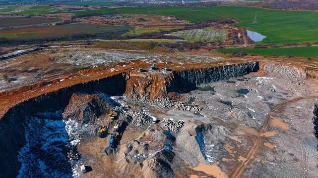 Mining equipment working on different levels of quarry. High angle shot of excavators and loaders moving stone in a deep granite pit near farm fields.