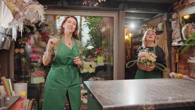 Two florists arranging bouquets in shop, green aprons at counter trimming stems and layering petals among vibrant blooms, warm lights and rustic shelves create inviting smallbusiness atmosphere