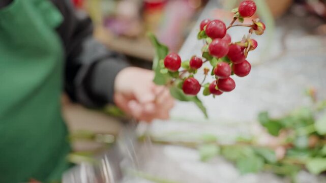 Florist trimming red berry stems, hands using shears removing leaves and aligning stems on workbench, green apron visible, rustic shop tabletop, focused calm atmosphere, seasonal centerpiece