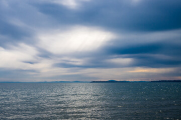 Obraz premium A dramatic, overcast sky with sunbeams breaking through the dark clouds illuminates the rippling surface of the sea, with distant hills visible on the horizon.
