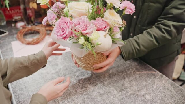 Florist presents pastel bouquet at counter, hands arrange pink and cream peonies and lisianthus in woven basket, customer reaches to inspect arrangement, wrapping sheet and wreath materials on table,