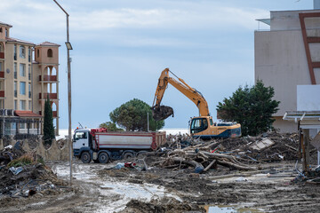 Fototapeta premium heavy machinery at work in the aftermath of flooding in Elenite, Bulgaria, where a yellow excavator is loading debris into a white dump truck on a mud-covered ground between resort buildings.