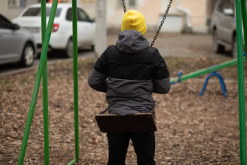 A child in a yellow beanie and dark winter jacket swings thoughtfully in an urban playground