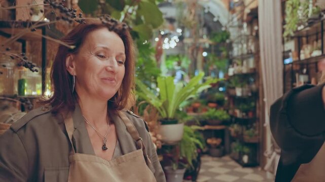 Florist shop consultation with plants, brunette florist in apron gives gentle guidance while touching stems and pointing to display shelves, boutique interior filled with pots and greenery, warm