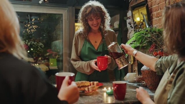 Florist team sharing tea at counter, trio of women gather around display of flowers and pastries, pouring tea into red mugs, warm string lights and window reflections, friendly small business