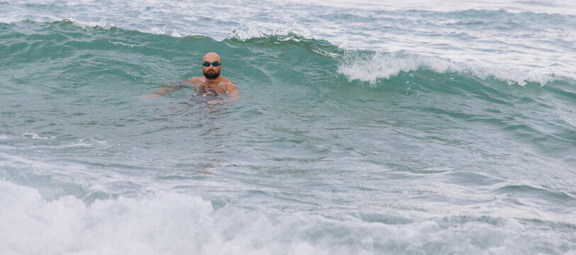 Bald man with beard swimming in rough ocean waves wearing goggles enjoying an outdoor water workout