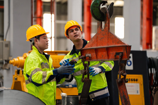 Two industrial workers in safety helmets inspecting lifting hook and chain system in factory, checking overhead crane equipment for maintenance and workplace safety compliance.